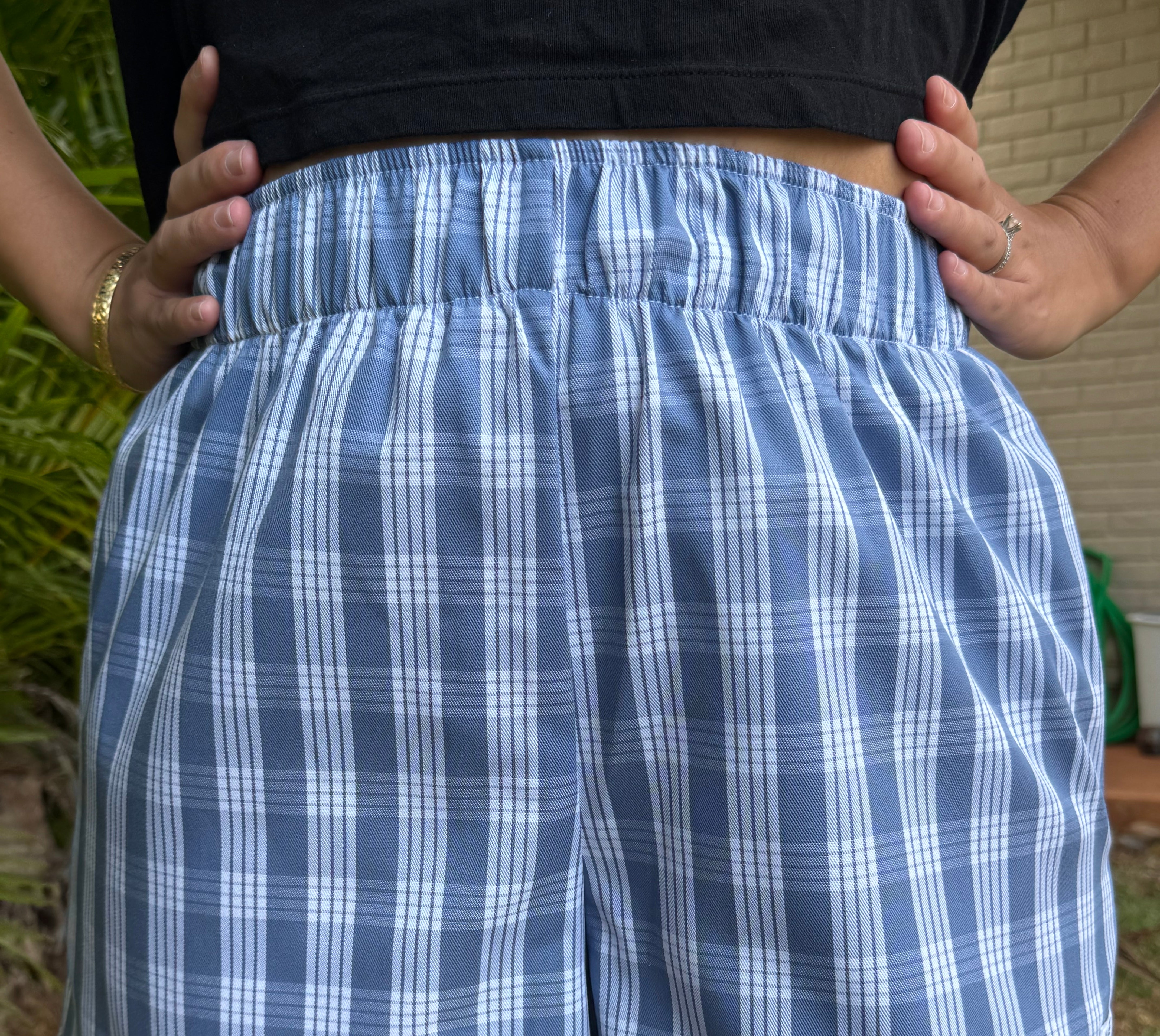 Wahine Holoholo Shorts 2.0 in Palaka print with elastic waist. Model's hands on hips, wearing a black top, outdoors in sunlight.