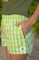 Wahine Holoholo Shorts 2.0 in a bright green Palaka pattern, featuring an elastic waistband. Hands are placed in pockets, paired with a green t-shirt.