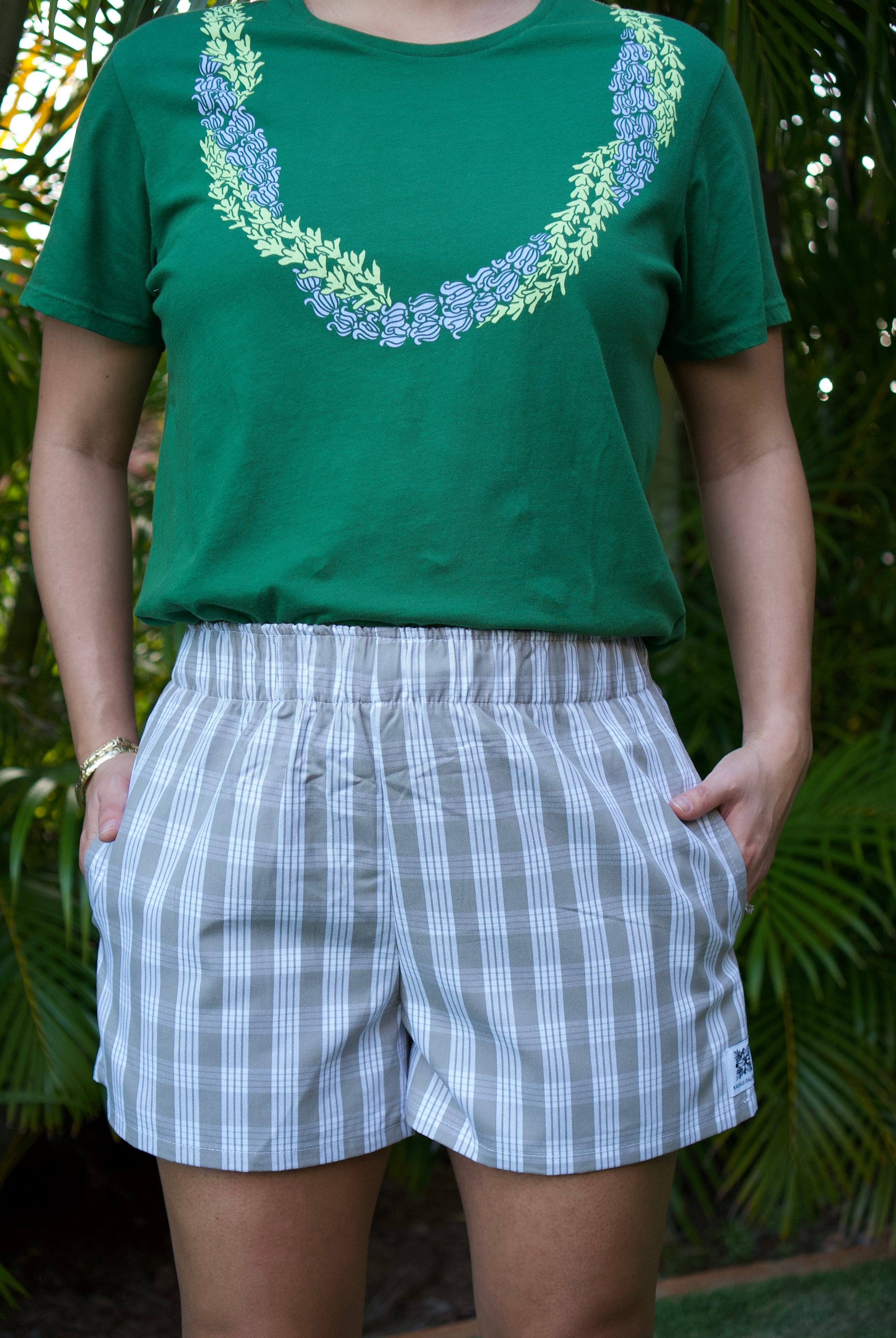Wahine Holoholo Shorts 2.0 feature a Palaka print with elastic waistband, worn with hands in pockets. Green shirt with floral design present.