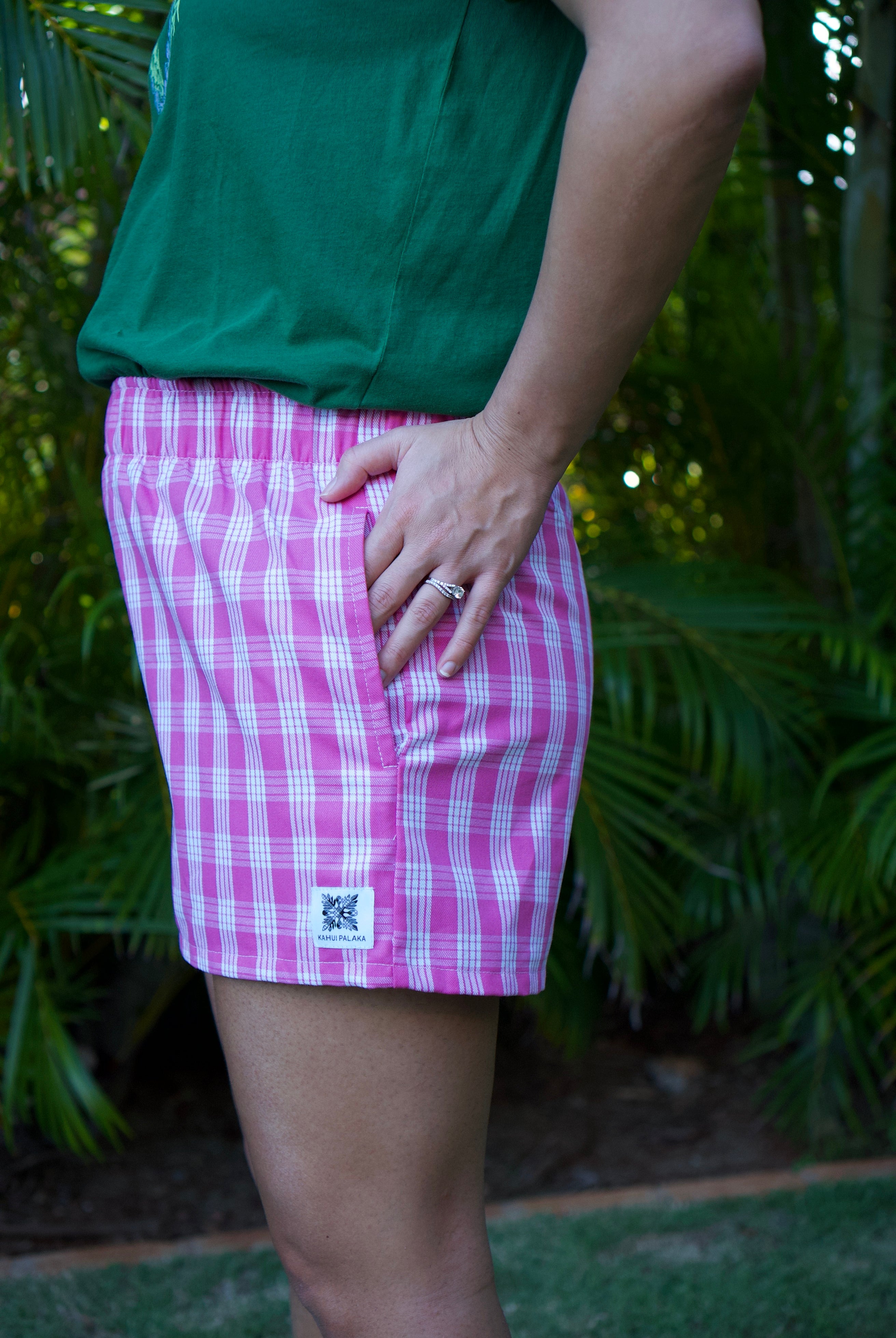 Wahine Holoholo Shorts 2.0 with traditional pink Palaka print. Side view shows pocket detail and logo patch. Outdoor setting.