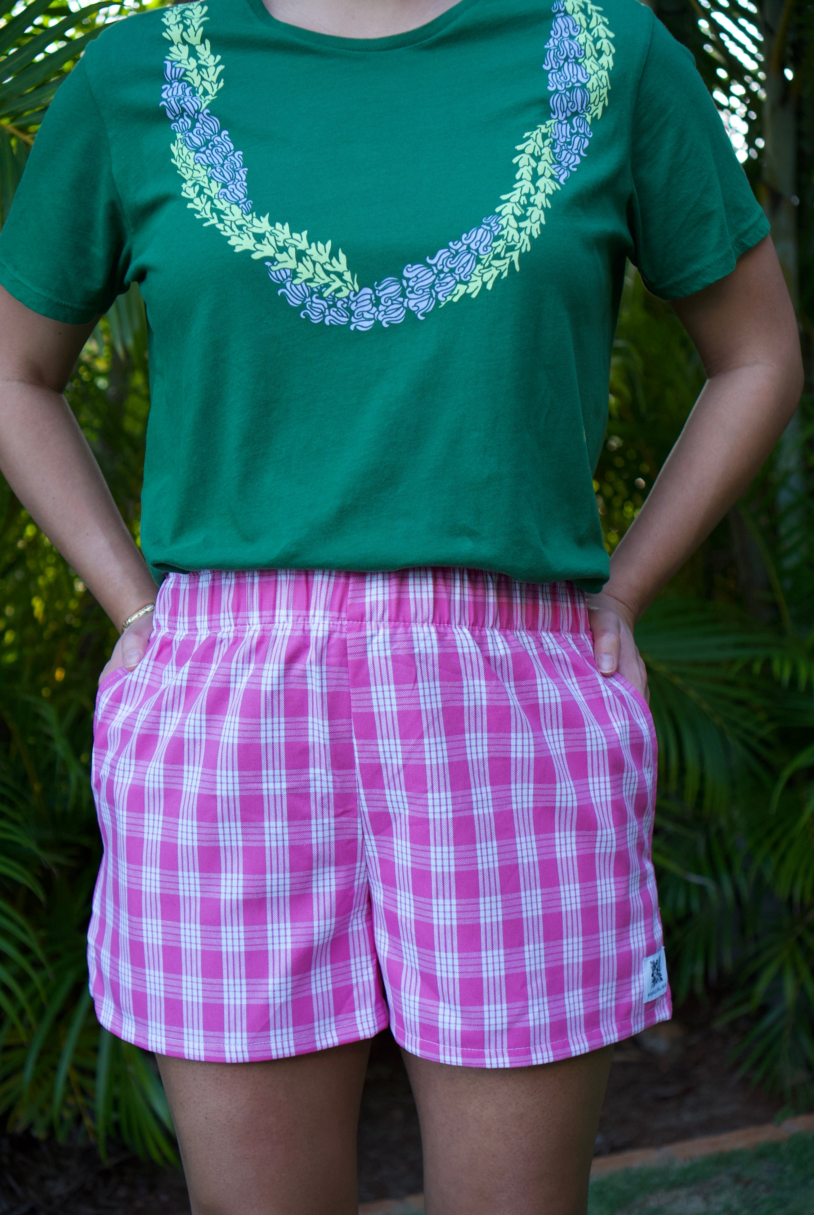 Lokelani Dark shorts with Palaka print, worn by someone posing with hands in pockets. Green tee with floral design.