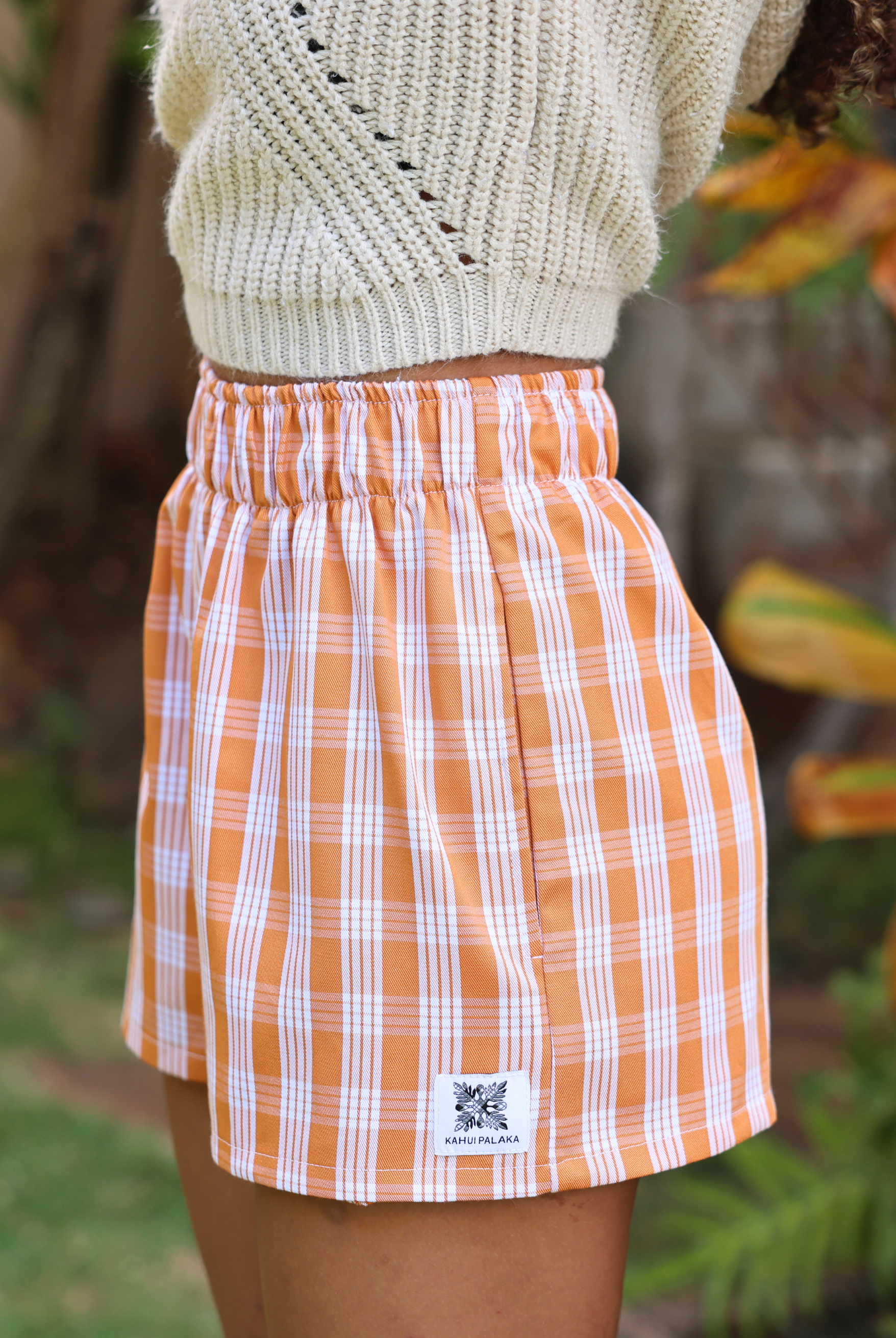 Wahine Holoholo Shorts 2.0 in Kaunaʻoa Rust with a traditional Palaka pattern. Elastic waistband, paired with a cream knit sweater.