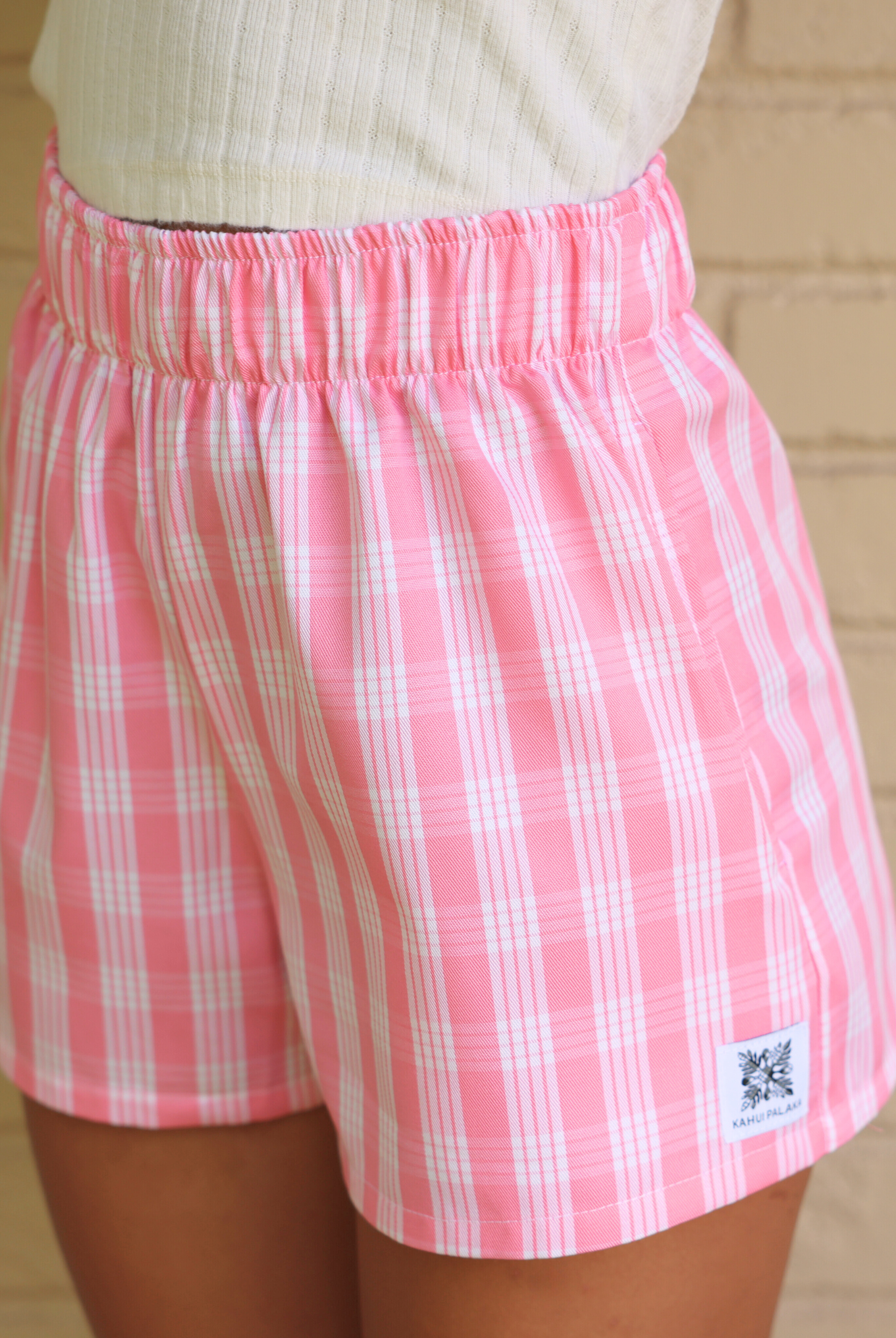 Wahine Holoholo Shorts 2.0 in Lokelani Light, featuring a pink Palaka pattern. Elastic waistband, side view, worn with a ribbed beige top.