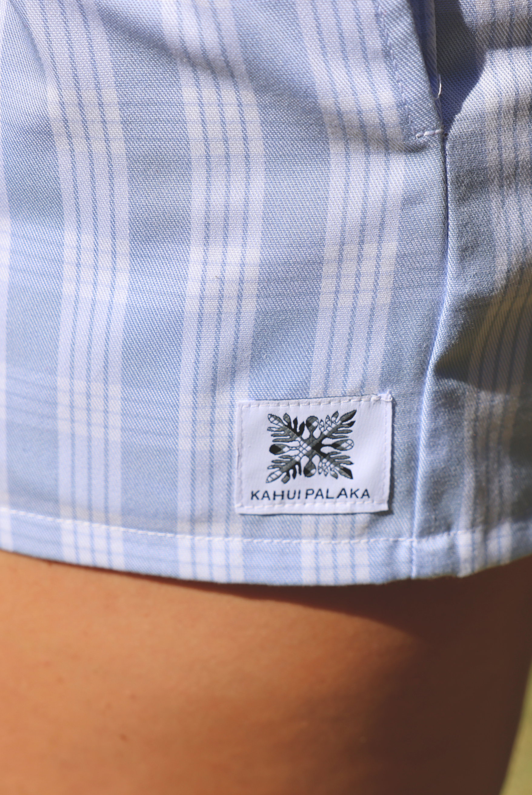 Wahine Holoholo Shorts 2.0 in light blue Palaka pattern. Close-up of side seam with brand tag. Sunlit, natural setting.