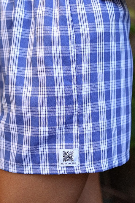 Wahine Holoholo Shorts feature an Imua Blue Palaka print. Side profile with garden plants in the blurred background.