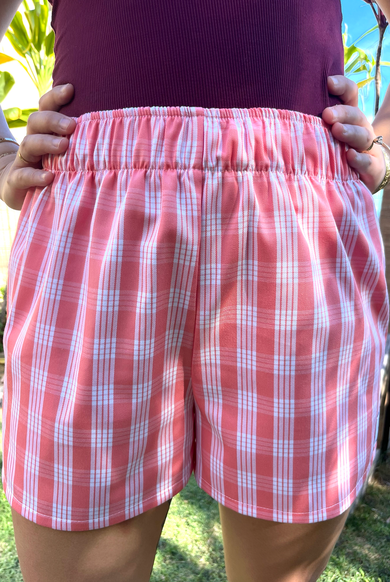 Wahine Holoholo Shorts 2.0 in ʻŌhelohelo feature a traditional Palaka print. Model poses with hands on hips. Bright sunlight highlights the shorts' pattern and colors.