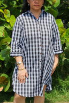 Moana Collection Dress features a traditional Palaka pattern with brown buttons. The model wears gold bracelets. Background has lush green leaves.