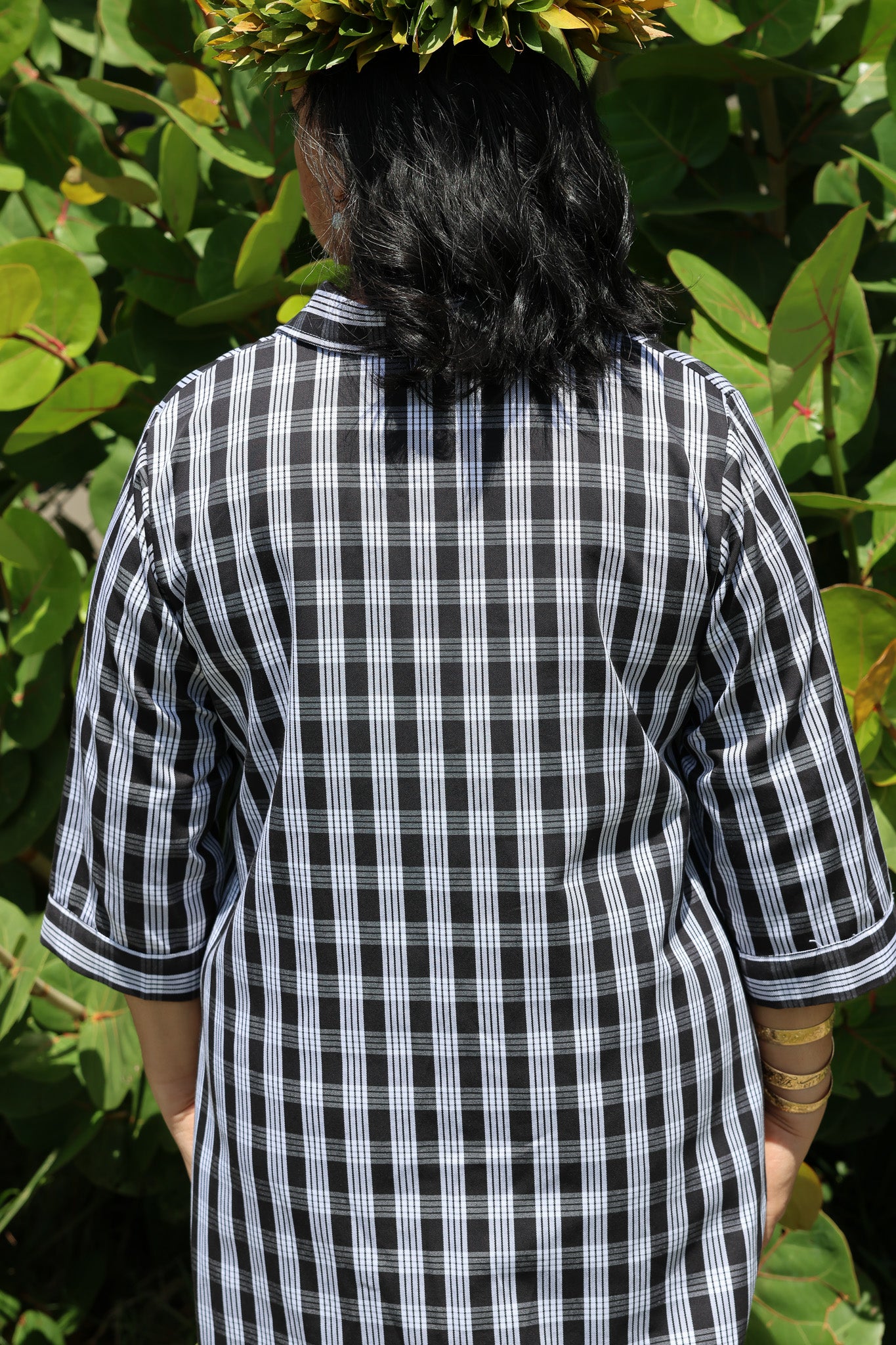 Back view of Moana Collection Dress in ʻAʻā with Palaka print. Model wears a leaf crown and gold bangles, standing outdoors.