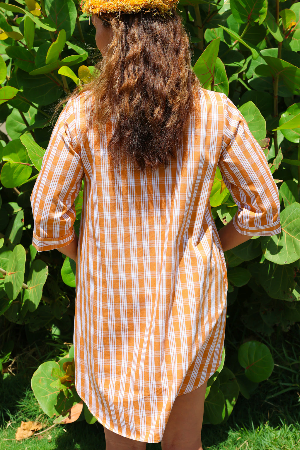 Dress in Kaunaʻoa Rust with Palaka print, viewed from the back. Features half sleeves. Model wears a lei, standing by lush greenery.