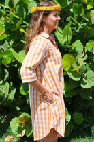 Kaunaʻoa Rust dress with Palaka print, mid-length sleeves. Model in side pose, hand in pocket, wearing a yellow head lei. Bright lighting with foliage background.
