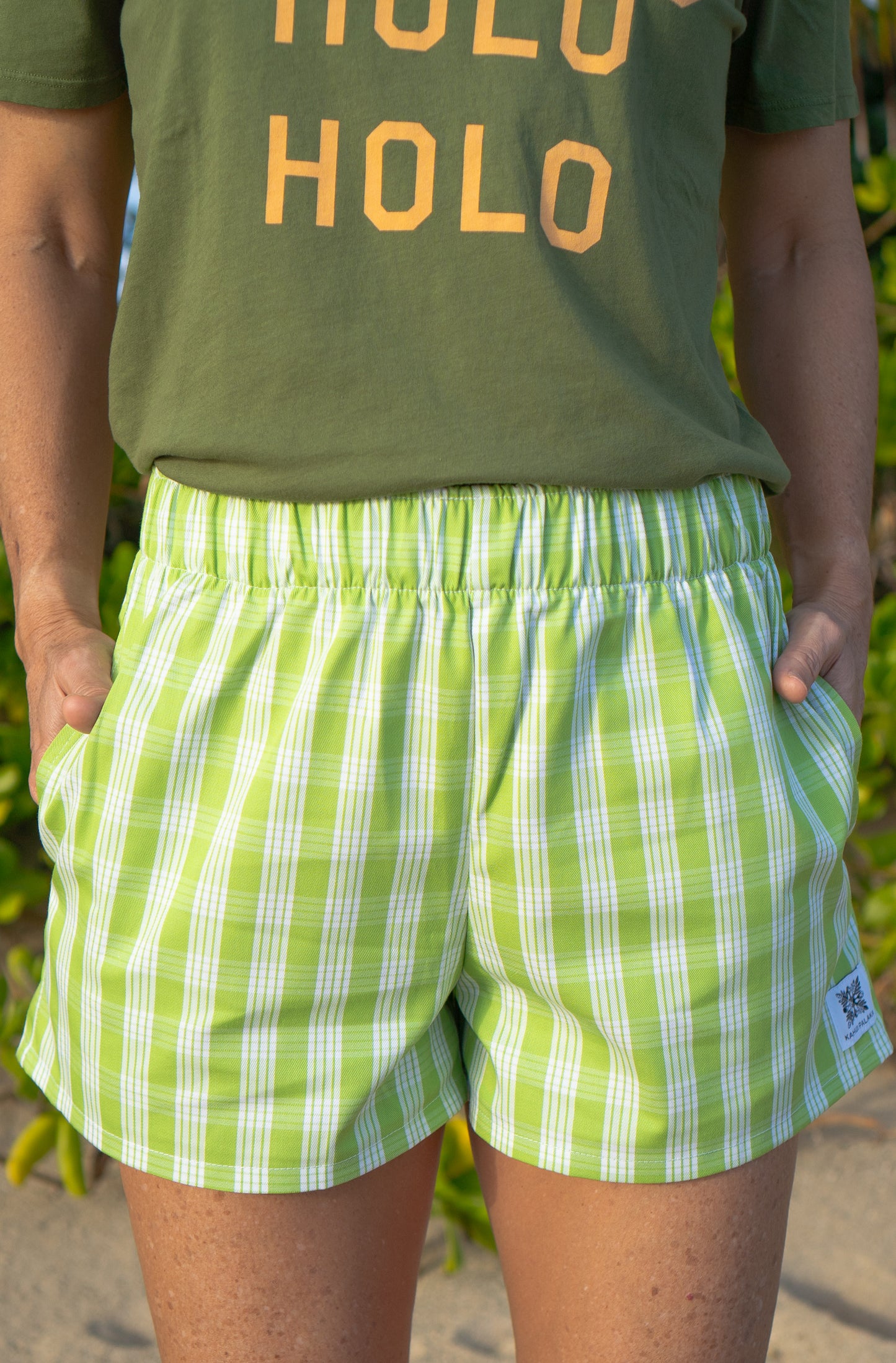 Wahine Holoholo Shorts 2.0 in a bright green Palaka pattern, featuring an elastic waistband. Hands are placed in pockets, paired with a green t-shirt.