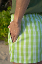 Wahine Holoholo Shorts 2.0 in a green Palaka pattern, shown from the side with hand in pocket, against a blurred nature background.