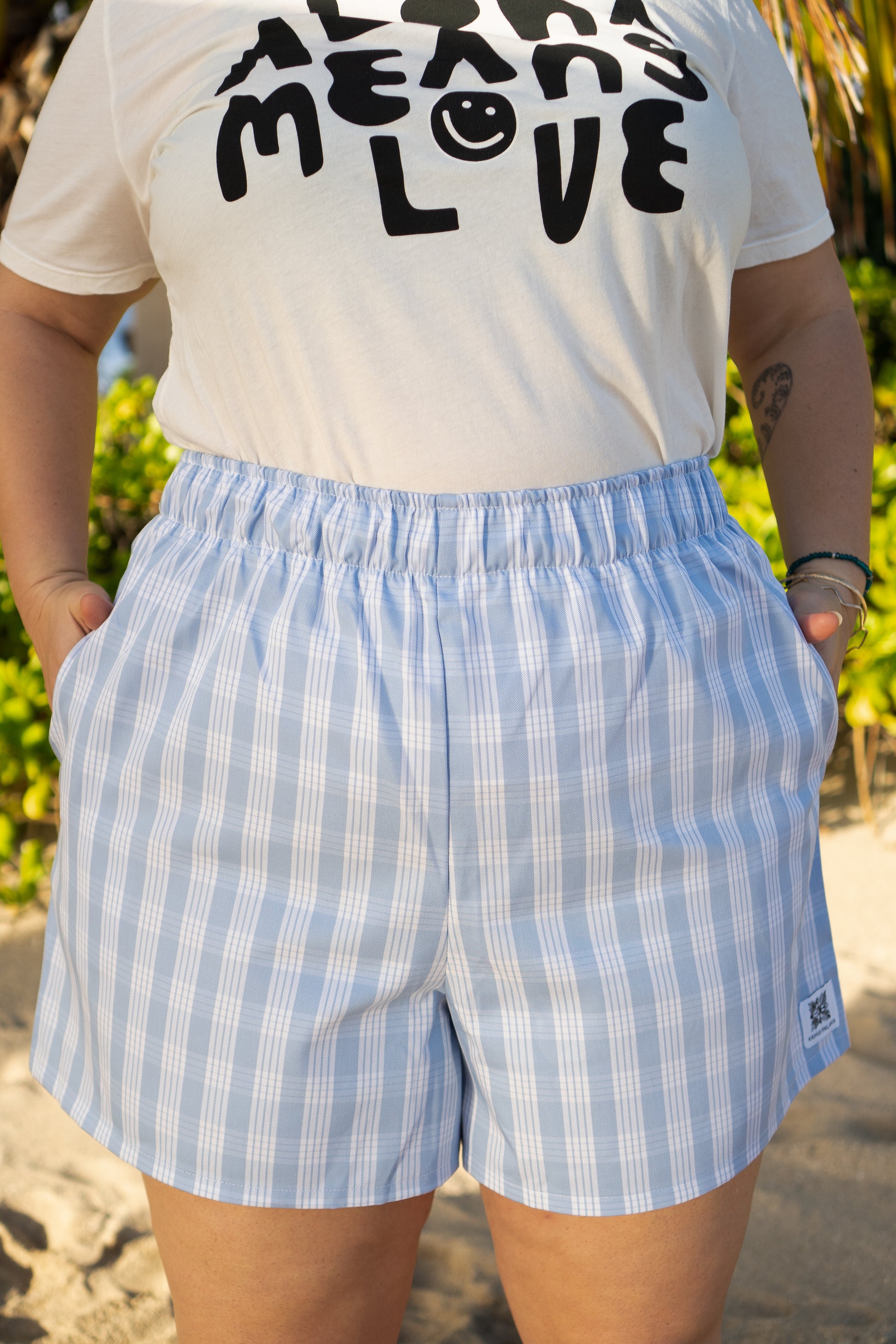 Wahine Holoholo Shorts 2.0 in ʻŌpua with a Palaka pattern. Model's hands in pockets, wearing a graphic tee. Bright outdoor setting.