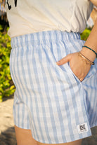 Wahine Holoholo Shorts 2.0 in soft blue Palaka print, elastic waistband, hand in pocket. Worn with a beige top, outdoors.