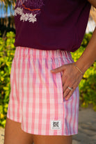 Wahine Holoholo Shorts 2.0 in Lokelani Light feature a pink Palaka pattern. Model stands with one hand in pocket, wearing rings and a bracelet.