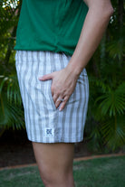 Wahine Holoholo Shorts 2.0 in Hinahina feature a traditional Palaka print. Modeled outdoors with a hand in the pocket, paired with a green top.