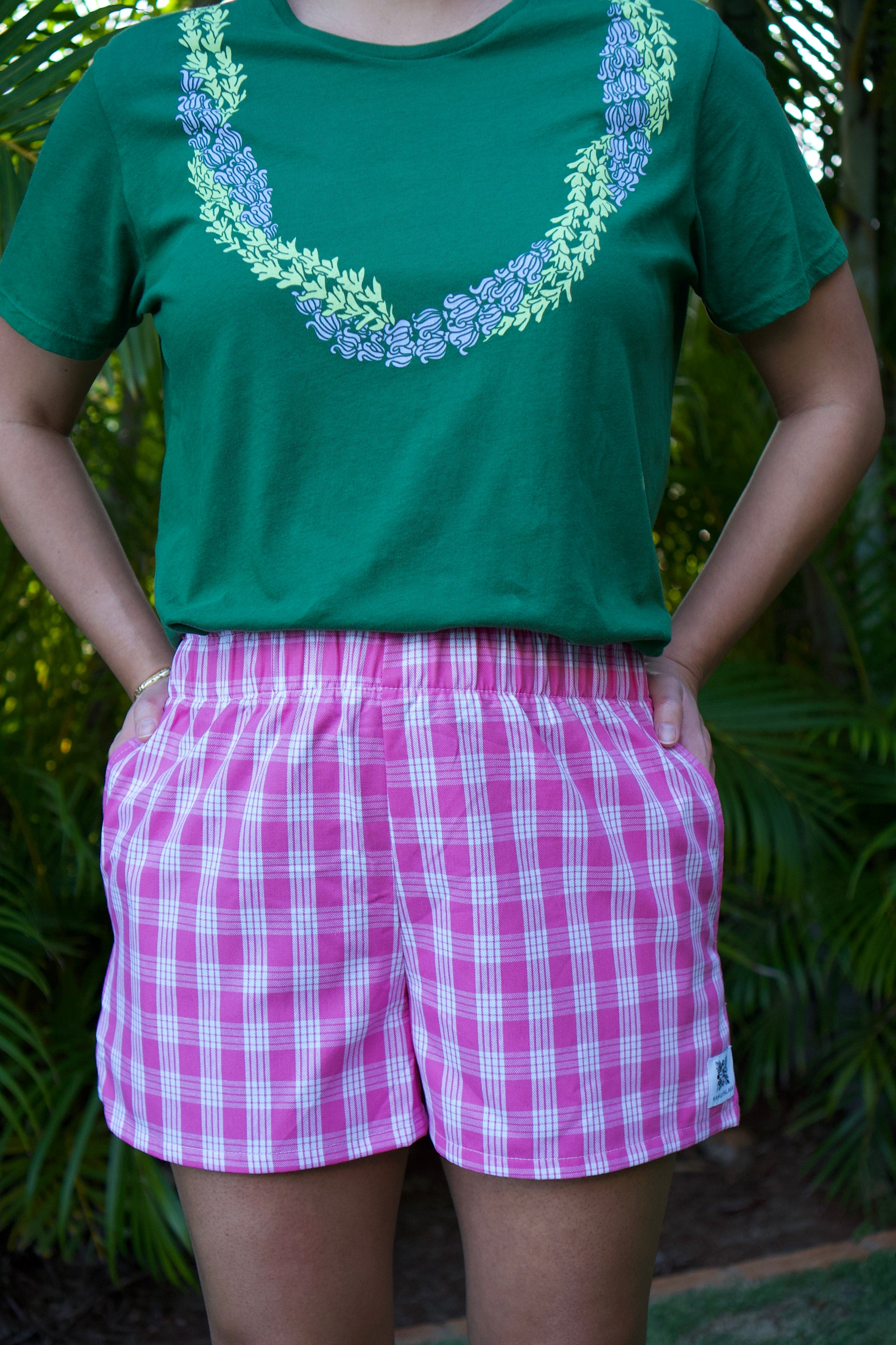 Lokelani Dark shorts with Palaka print, worn by someone posing with hands in pockets. Green tee with floral design.