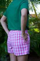 Wahine Holoholo Shorts 2.0 in Lokelani Dark, featuring a pink Palaka pattern. Model stands outdoors, one hand in pocket, wearing gold bangles.