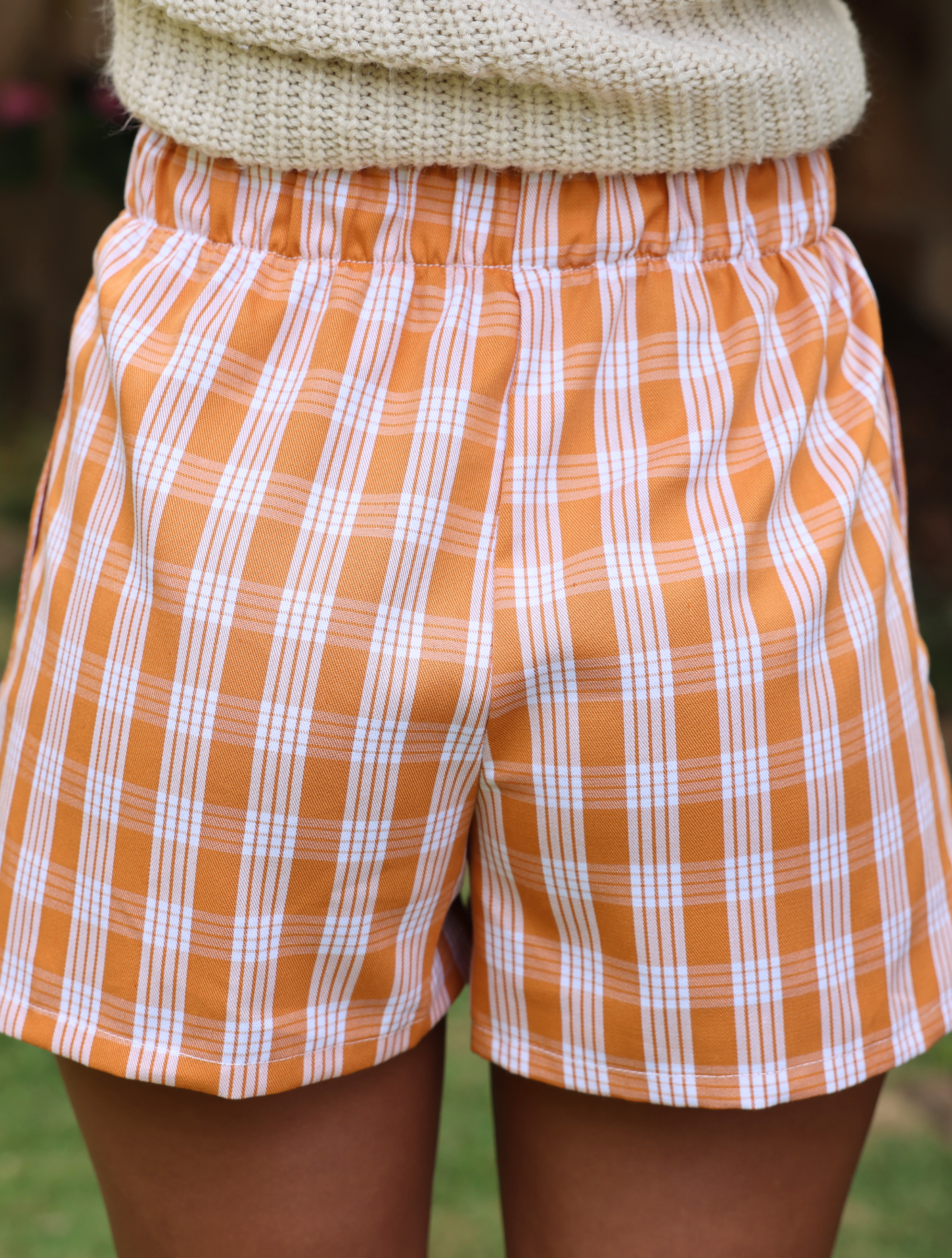 Wahine Holoholo Shorts 2.0 in Kaunaʻoa Rust Palaka print. Elastic waistband, paired with a beige knit top, outdoor setting.