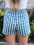 Back view of Wahine Holoholo Shorts 2.0 in Uluwehi, featuring green Palaka print. Elastic waistband, outdoors background.