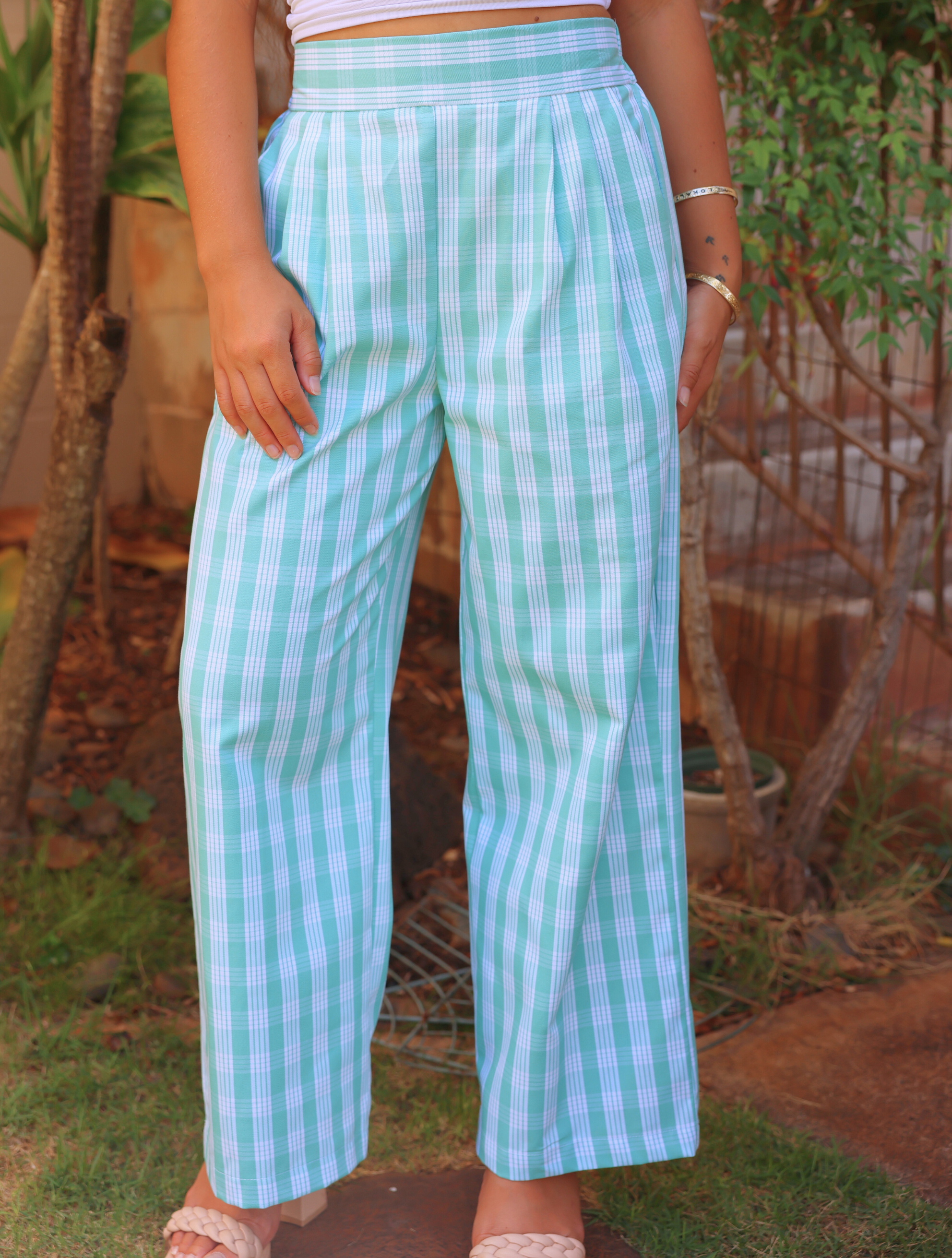 Liʻi Pants with a light blue and white Palaka print, worn by a model outdoors. High-waisted with pleats, paired with a white top and braided sandals.