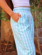 Liʻi Pants in a light blue Palaka print are shown in side profile. A hand rests in the pocket, worn with a white, ruched crop top. Background features greenery and warm lighting.