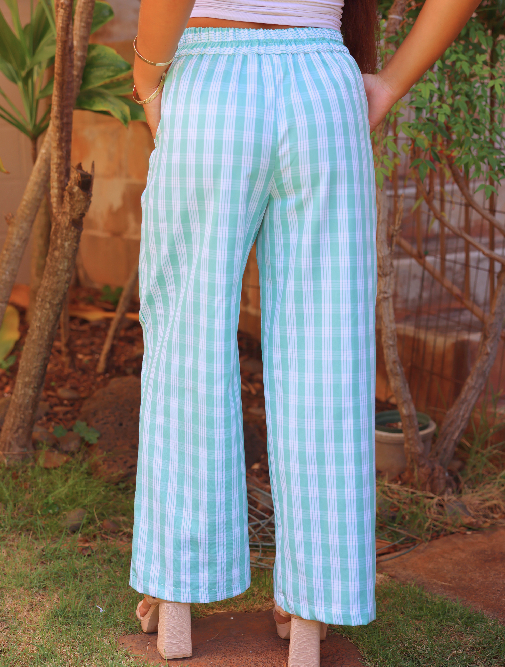 Liʻi Pants with traditional Palaka print, shown from the back. Elastic waistband, wide leg fit. Model outdoors, wearing beige heels and bangles.