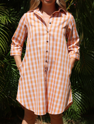 Kaunaʻoa Rust dress with Palaka print, button-front, and roll-up sleeves. Posed with hands in pockets, foliage background.