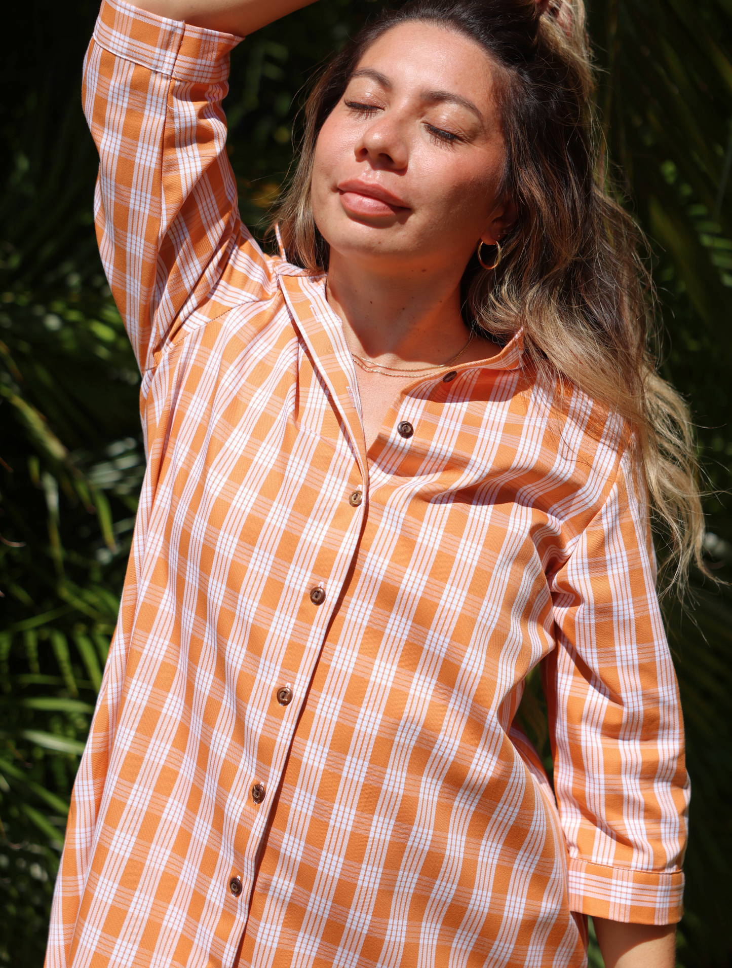 Moana Collection Dress in Kaunaʻoa Rust features a Palaka print. The dress has three-quarter sleeves and wooden buttons. Model poses with eyes closed, hand raised, wearing hoop earrings in bright sunlight.