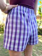 Wahine Holoholo Shorts 2.0 in purple Palaka print with elastic waist. Model poses outdoors, one hand on hip, wearing a gold bracelet.