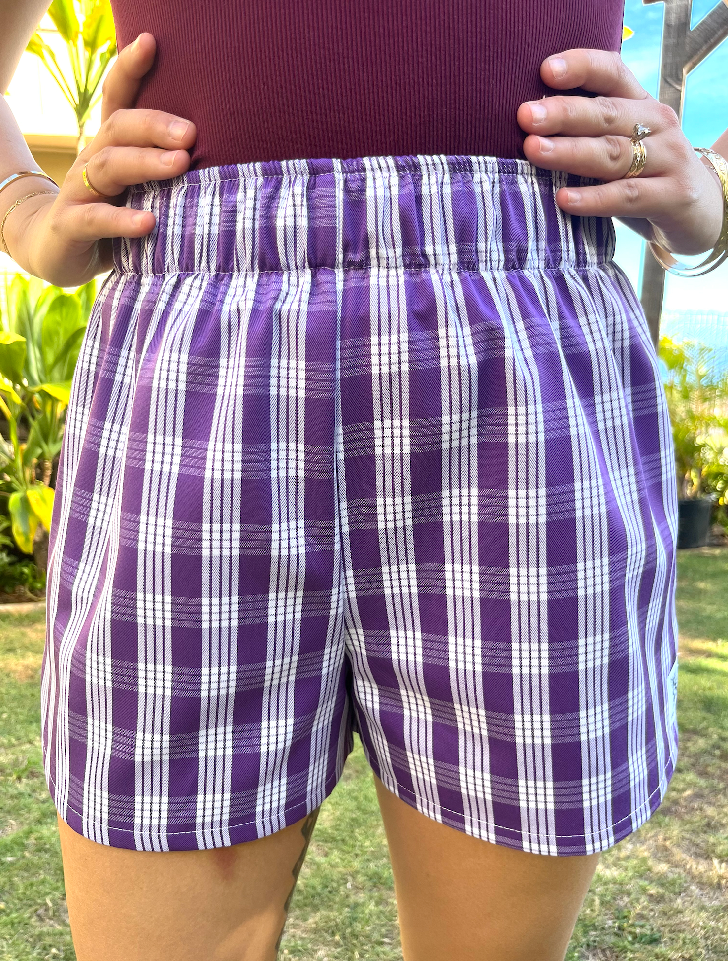 Wahine Holoholo Shorts 2.0 with Pōhuehue Palaka print, shown in bright outdoor light. Model holds waistband, showcasing fit.